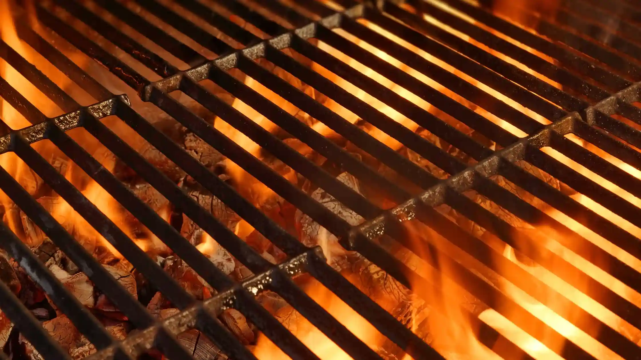 Glowing charcoal and bright flames burning beneath heavy cast-iron grill grates, emphasizing the importance of fire safety during summer cookouts.