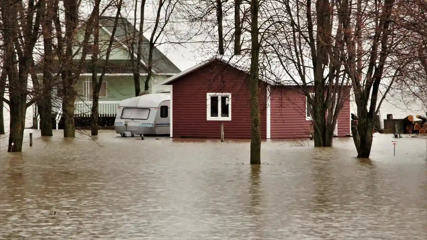 Residential properties surrounded by rising murky floodwaters that submerge the lower portions of the homes and nearby trees.