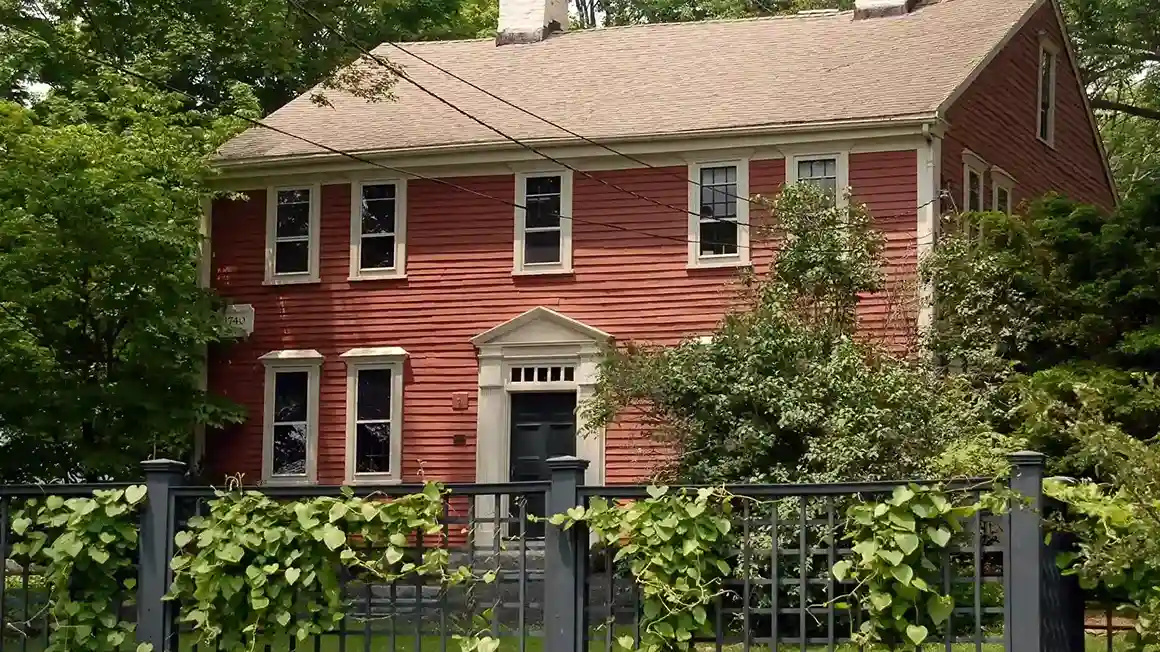 Traditional two-story colonial home with reddish siding, white trim, and a vine-covered fence, highlighting vacation season preparation for homeowners.