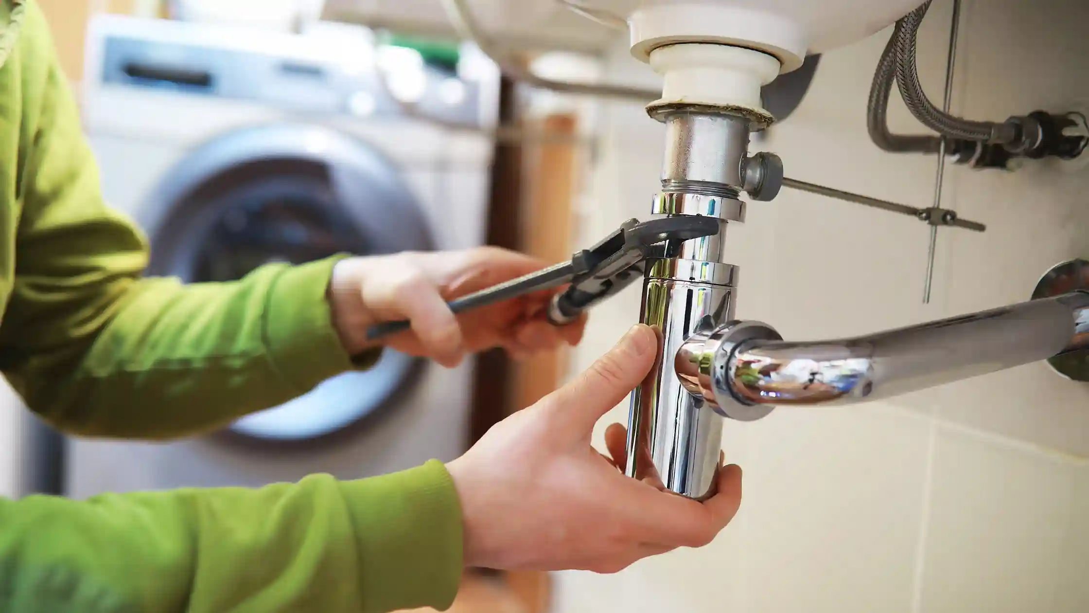 Person tightening sink plumbing with a wrench beneath a kitchen sink, supporting guidance on common summer plumbing issues for Massachusetts homeowners.