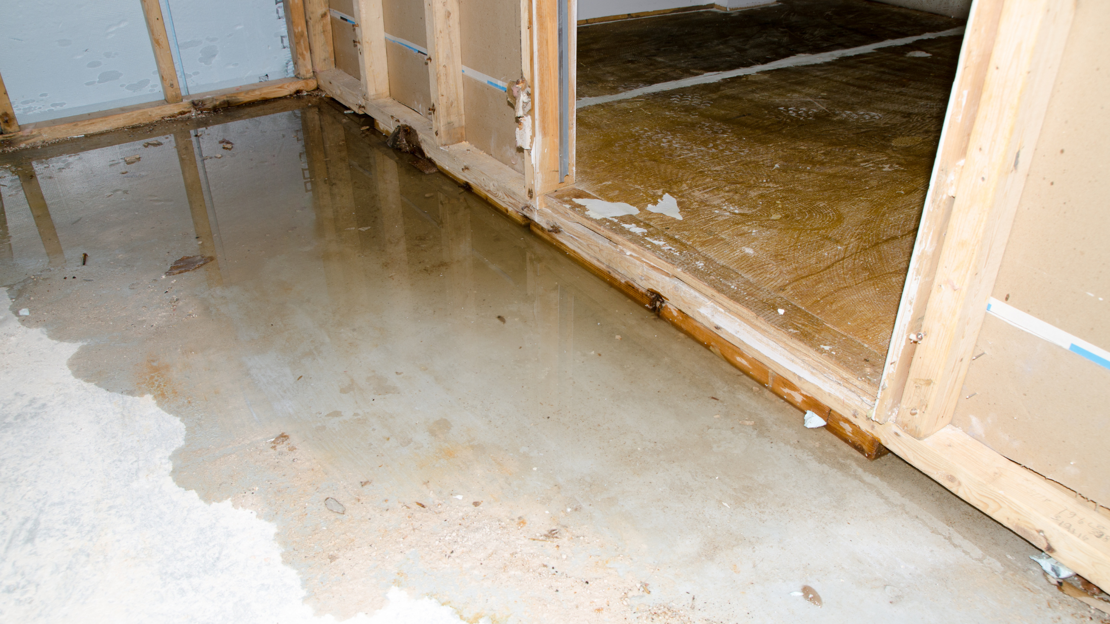Standing water covers the concrete floor of an unfinished room with exposed wooden framing and drywall partitions, showing significant indoor water damage.