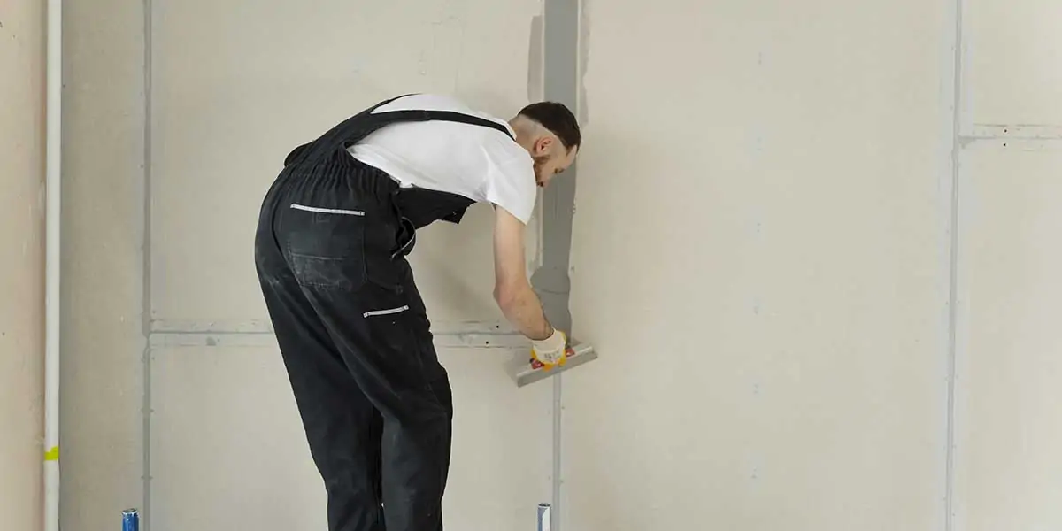 Worker in black overalls smoothing joint compound on a drywall seam, repairing wall damage to replace affected drywall panels.